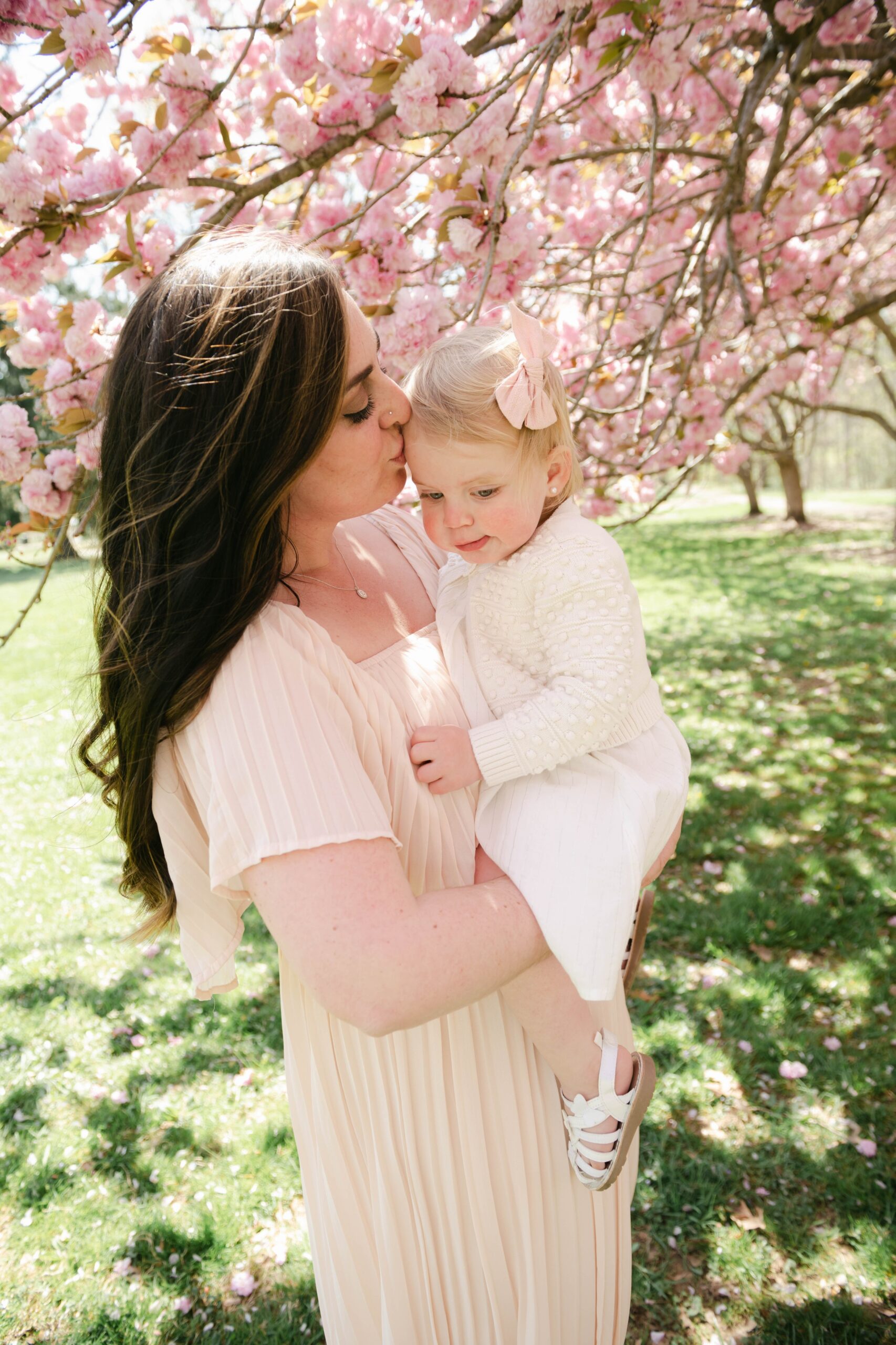 A mother in a beige dress kisses her toddler daughter's forehead while standing under pink tree blossoms luxey little ones