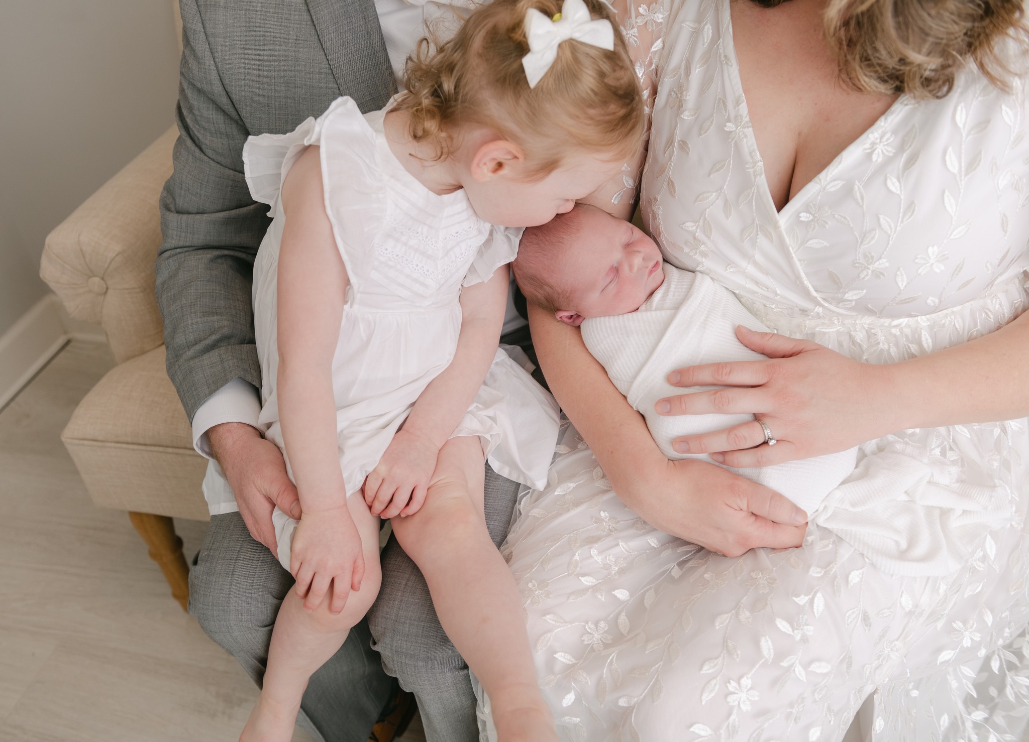 A mother in a white dress sits on a couch with her husband while she holds their newborn baby and their young daughter kisses the baby forehead chester county pediatricians
