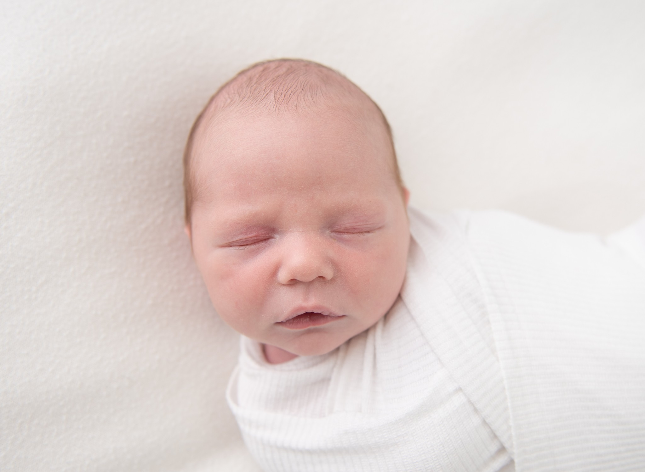 A newborn baby sleeps in a white swaddle on a white bed
