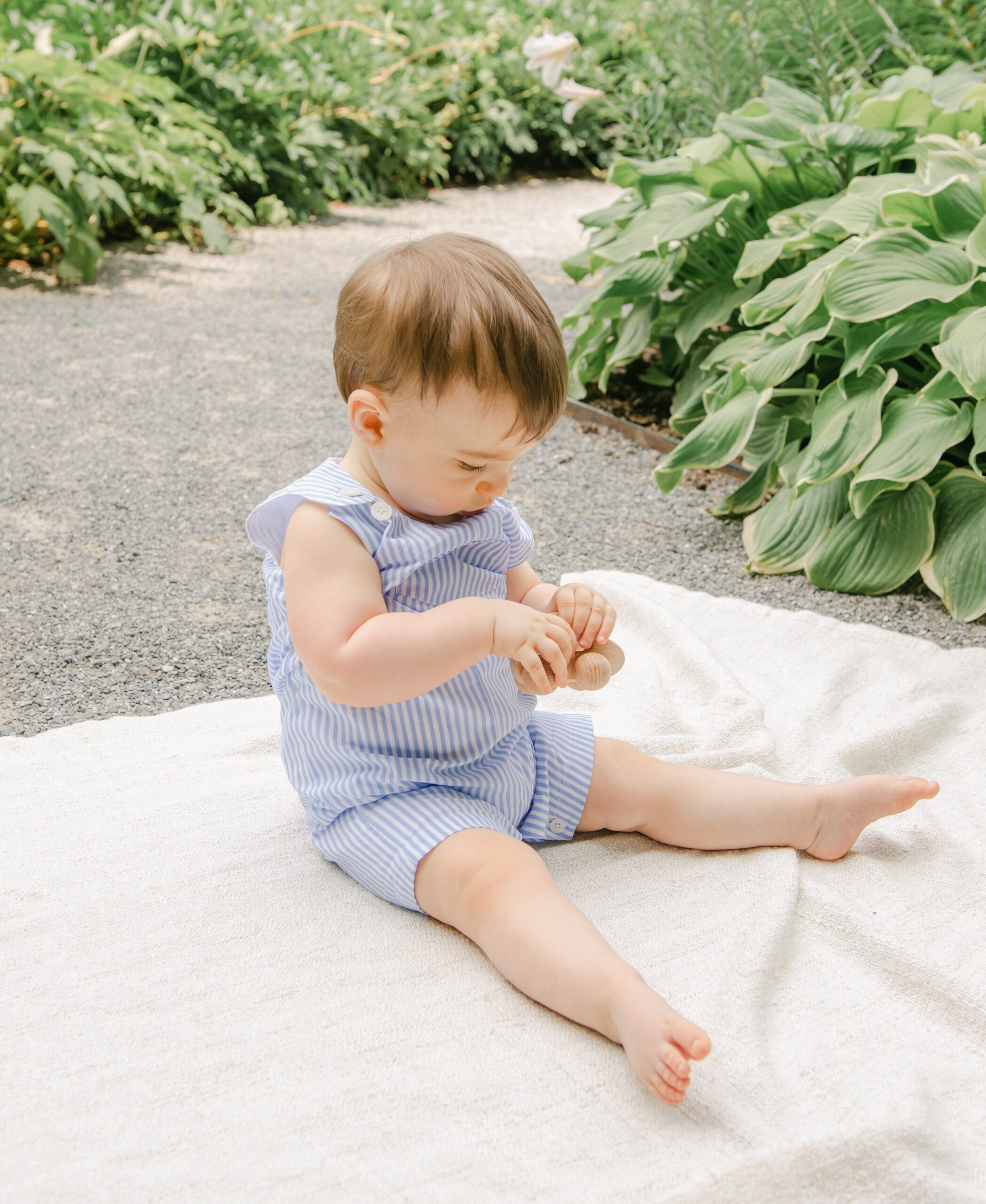 An infant boy sits on a blanket in a gravel garden path playing with a wooden toy