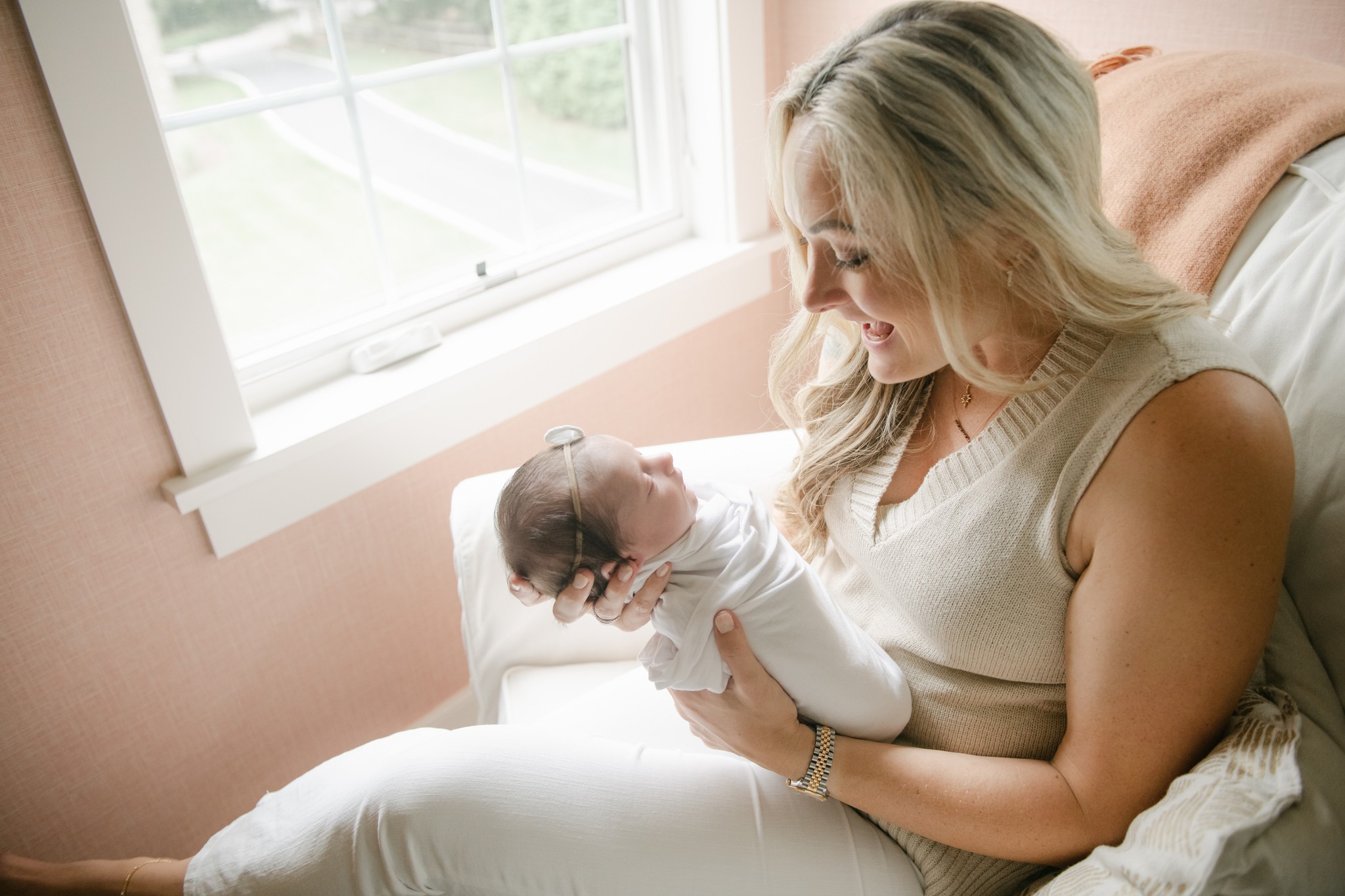 A new mom sits in a nursery chair in a pink room by a window smiling down at her newborn baby in her hands downington obgyn