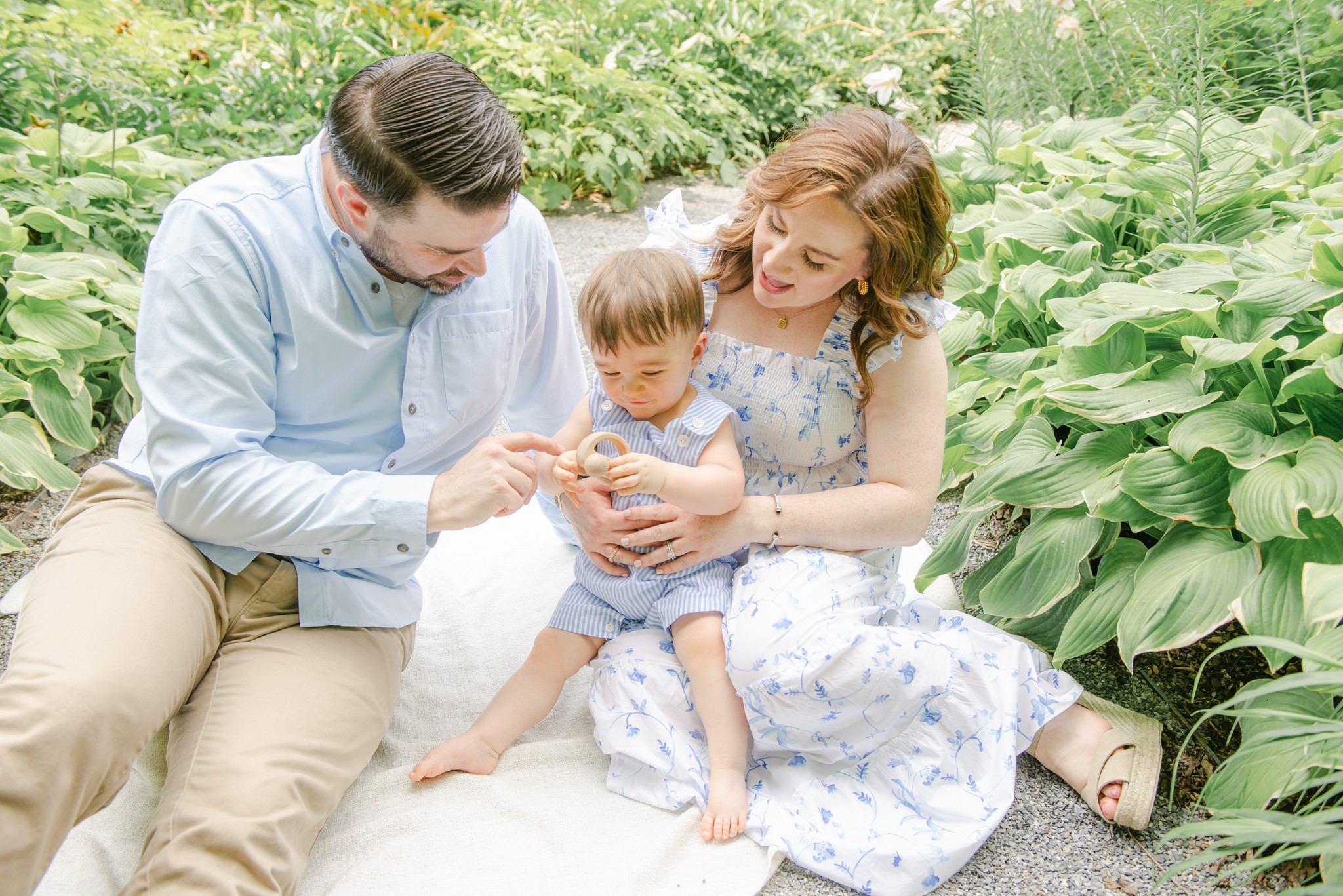A mother and father sit on a blanket in a gravel garden path playing with their infant son in mom's lap