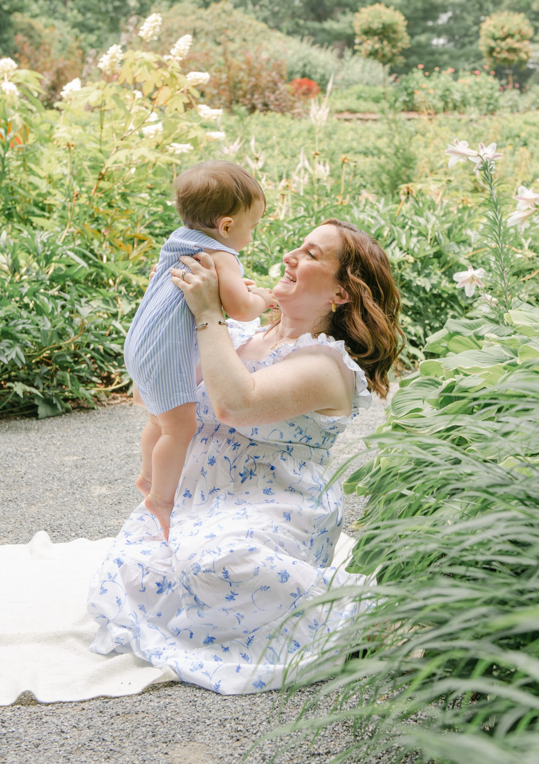 A mother in a white dress sits on a picnic blanket in a flower garden playing and lifting her infant son in a blue striped onesie downington preschools