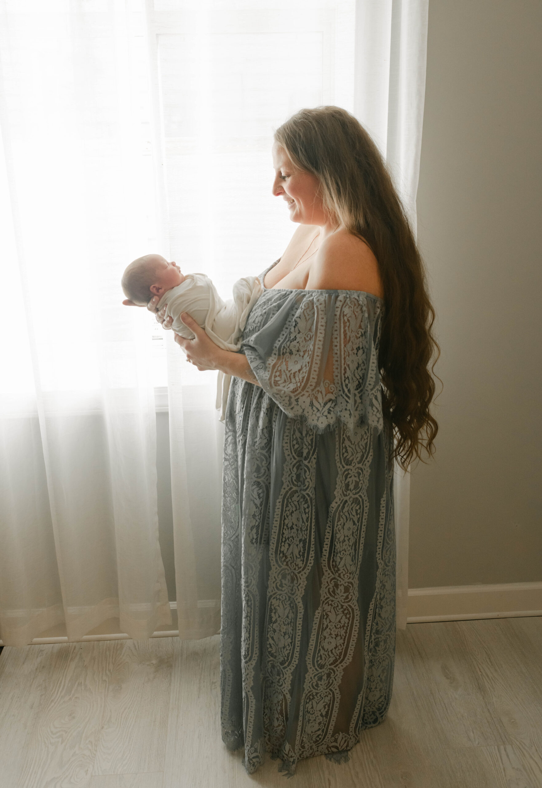A new mom wearing a long blue dress looks down at her sleeping newborn baby in front of a window in her hands