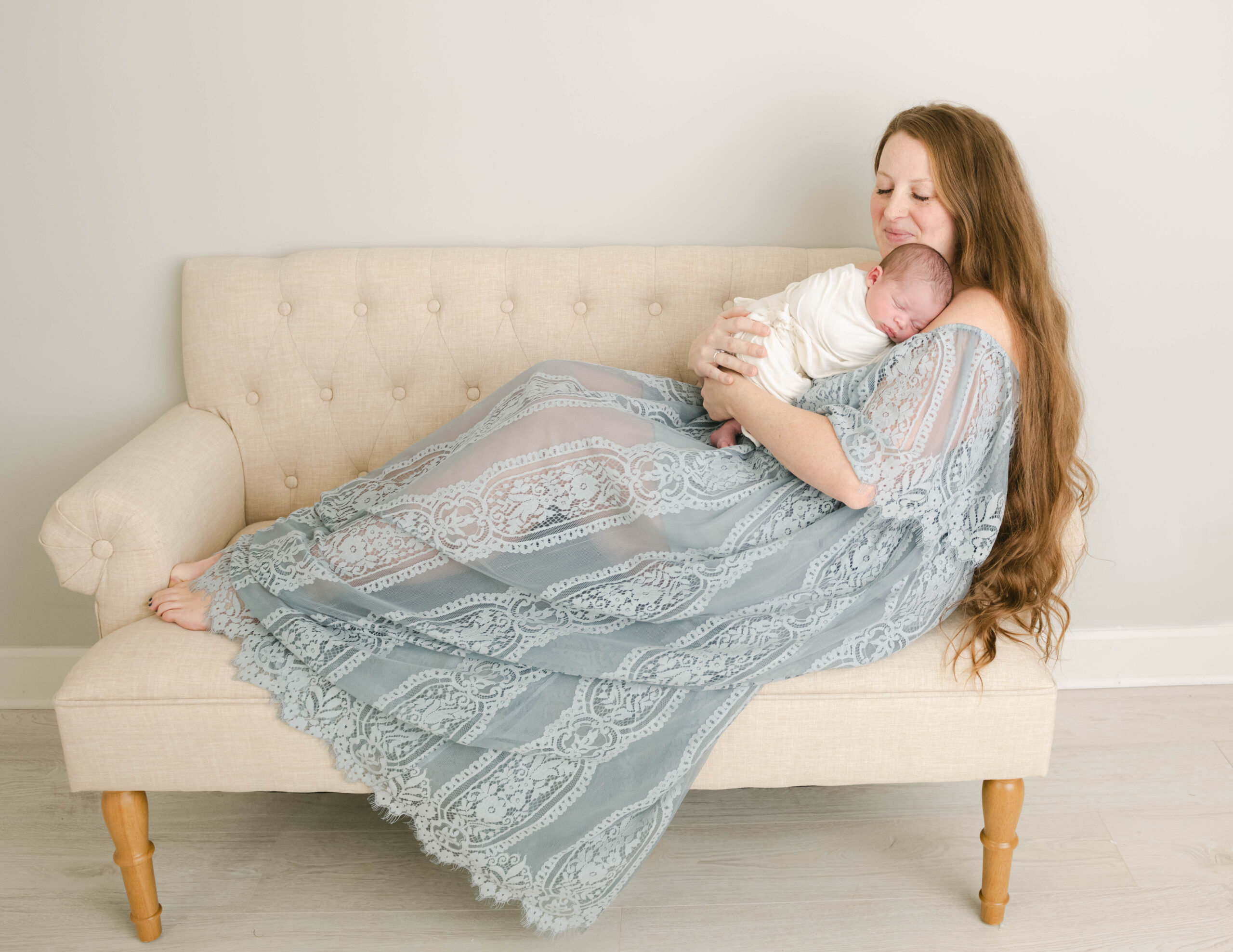 A new mom sits across a couch in a blue dress with her newborn baby sleeping on her shoulder prenatal yoga west chester pa
