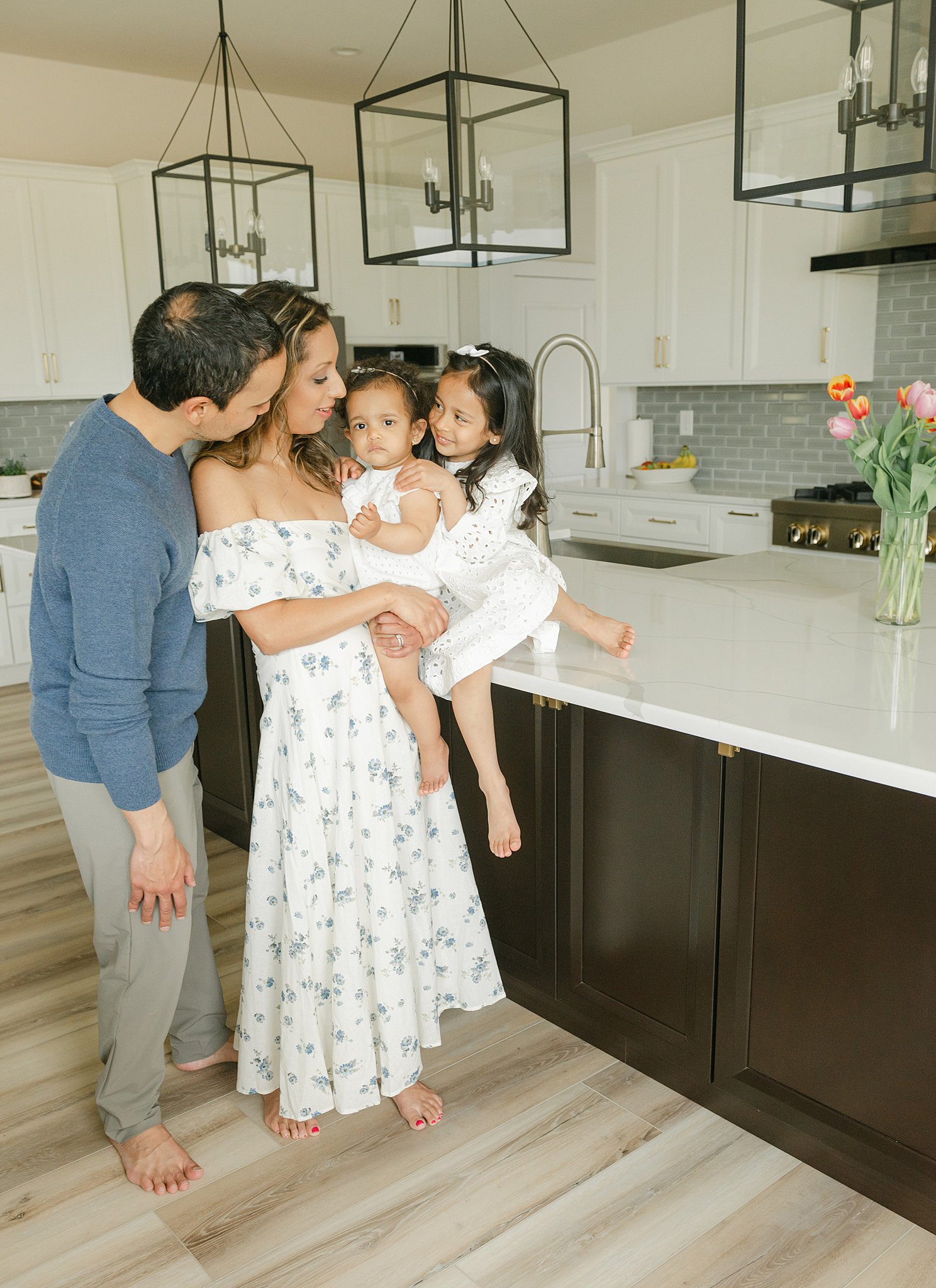 A mom holds her infant daughter while dad and their young daughter lean in on either side in a kitchen