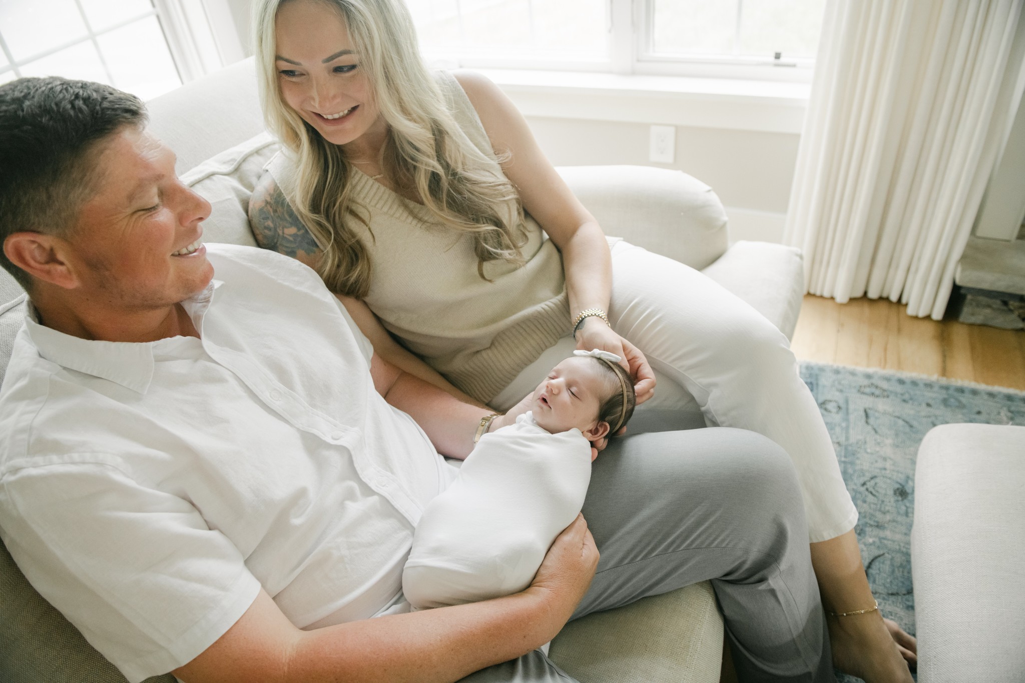 A mom and dad smile down at their sleeping newborn baby in dad's lap as they sit on a couch under a window