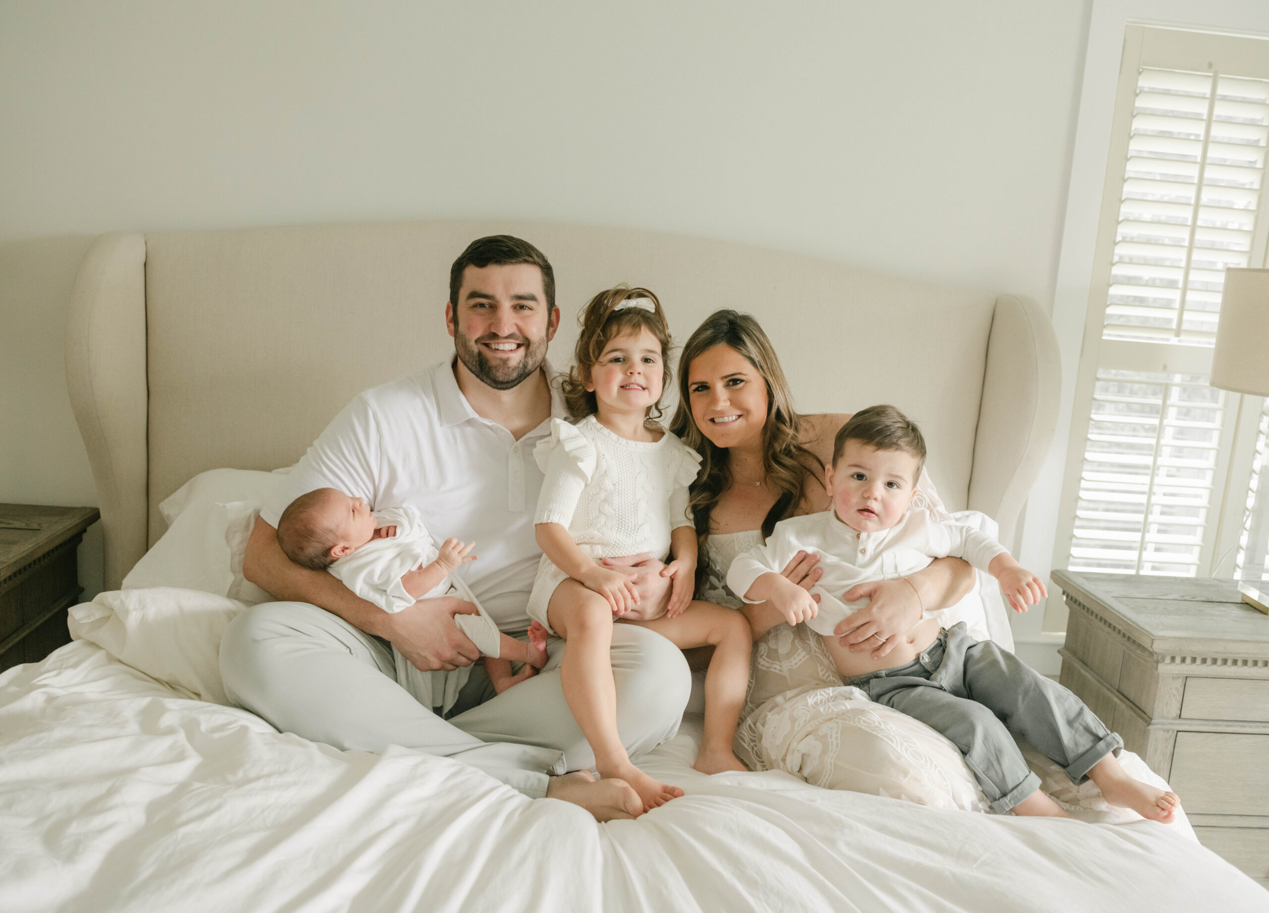 A mom and dad sit on a white bed with their two toddler children in their laps and dad holds their newborn baby in one arm maternal fetal medicine west chester