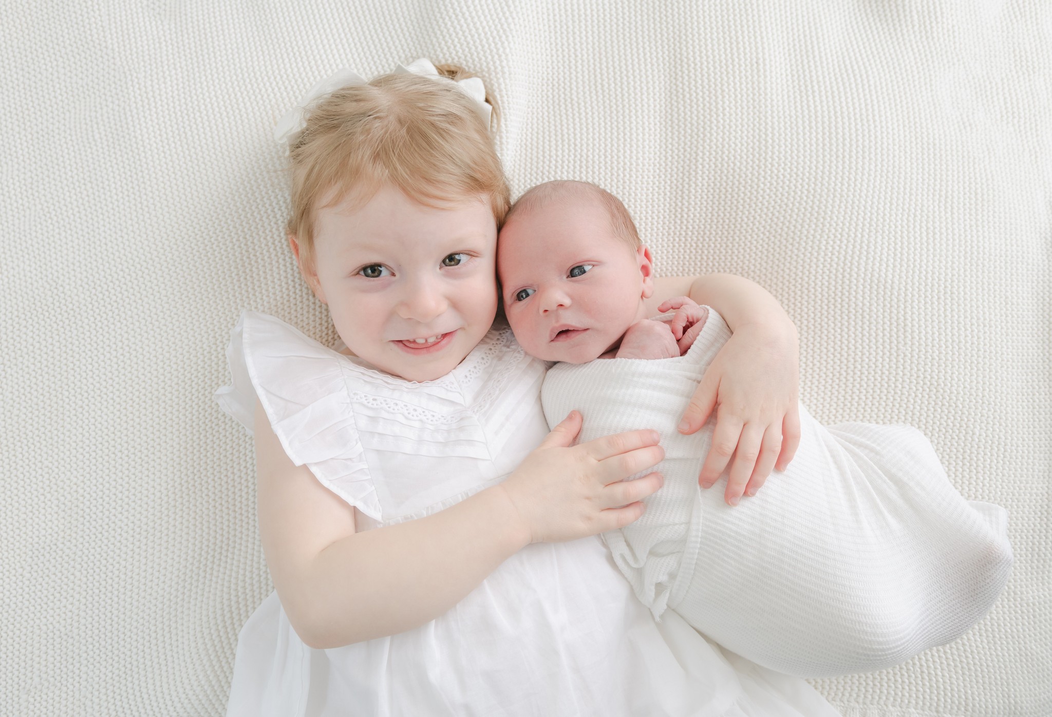 A toddler girl in a white dress cuddles on a white bed with her newborn baby sibling in a white swaddle