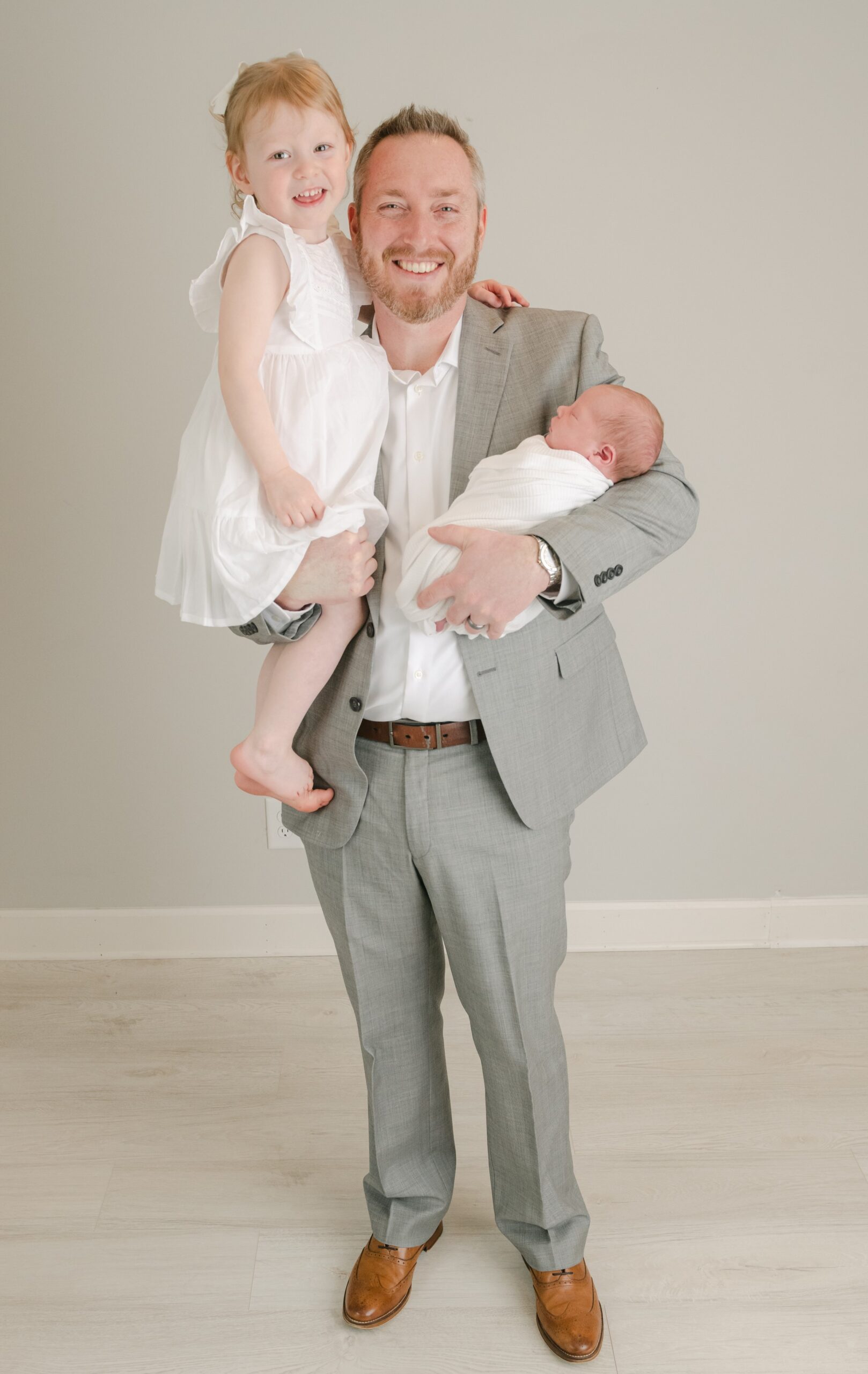 A happy father in a grey suit stands in a studio with his toddler daughter on one hip and his sleeping newborn baby in his other arm well born baby