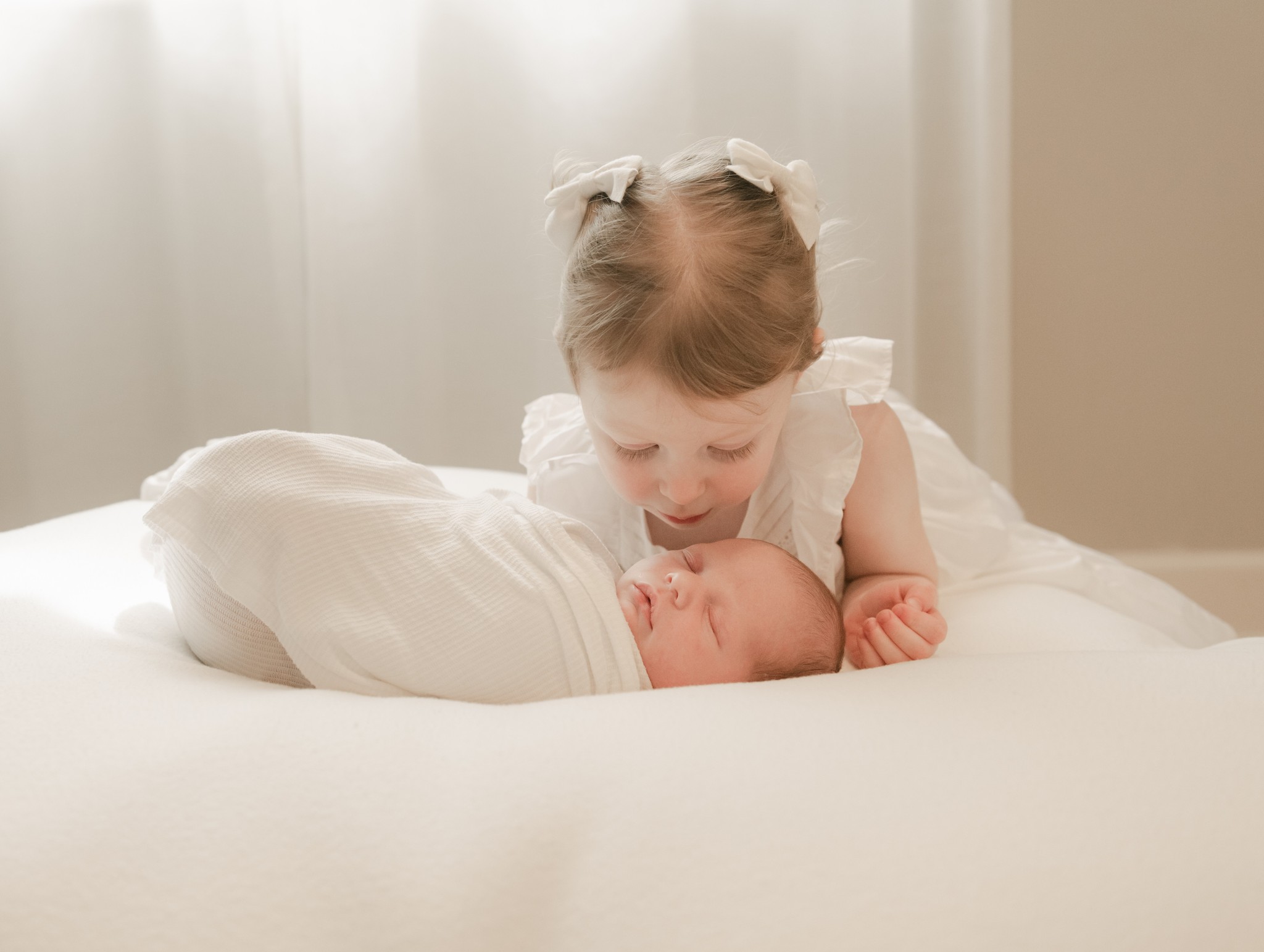 A toddler girl in a white dress lays on a bed overtop her sleeping newborn baby sibling well born baby