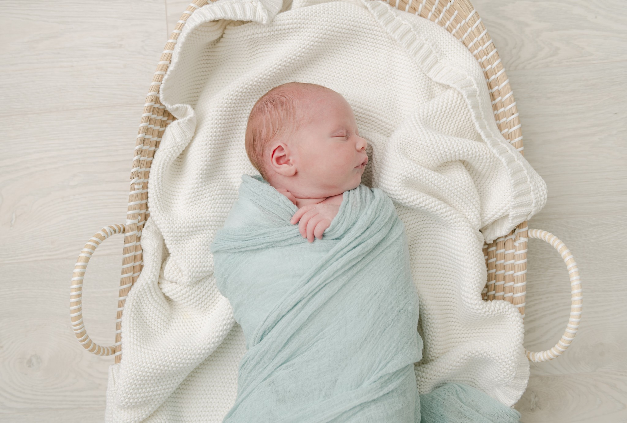 A newborn baby sleeps in a woven basket and green swaddle