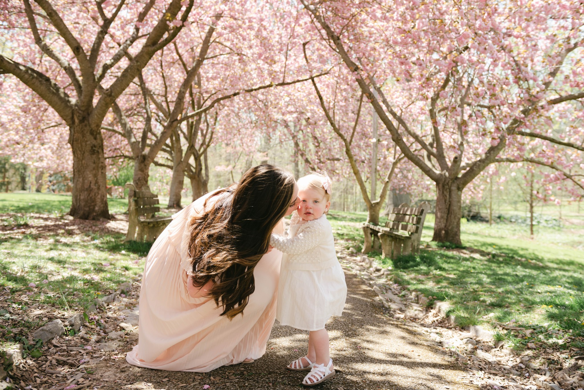A mom in a pink dress kneels down to kiss her toddler daughter in a white dress in a park path lined with pink blossoming trees from layla's boutique