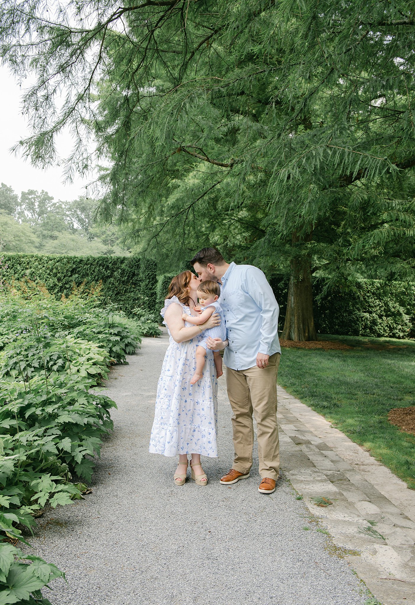 A mom and dad kiss while standing in a garden path and holding their toddler son all in blue and white because of a nanny lancaster pa