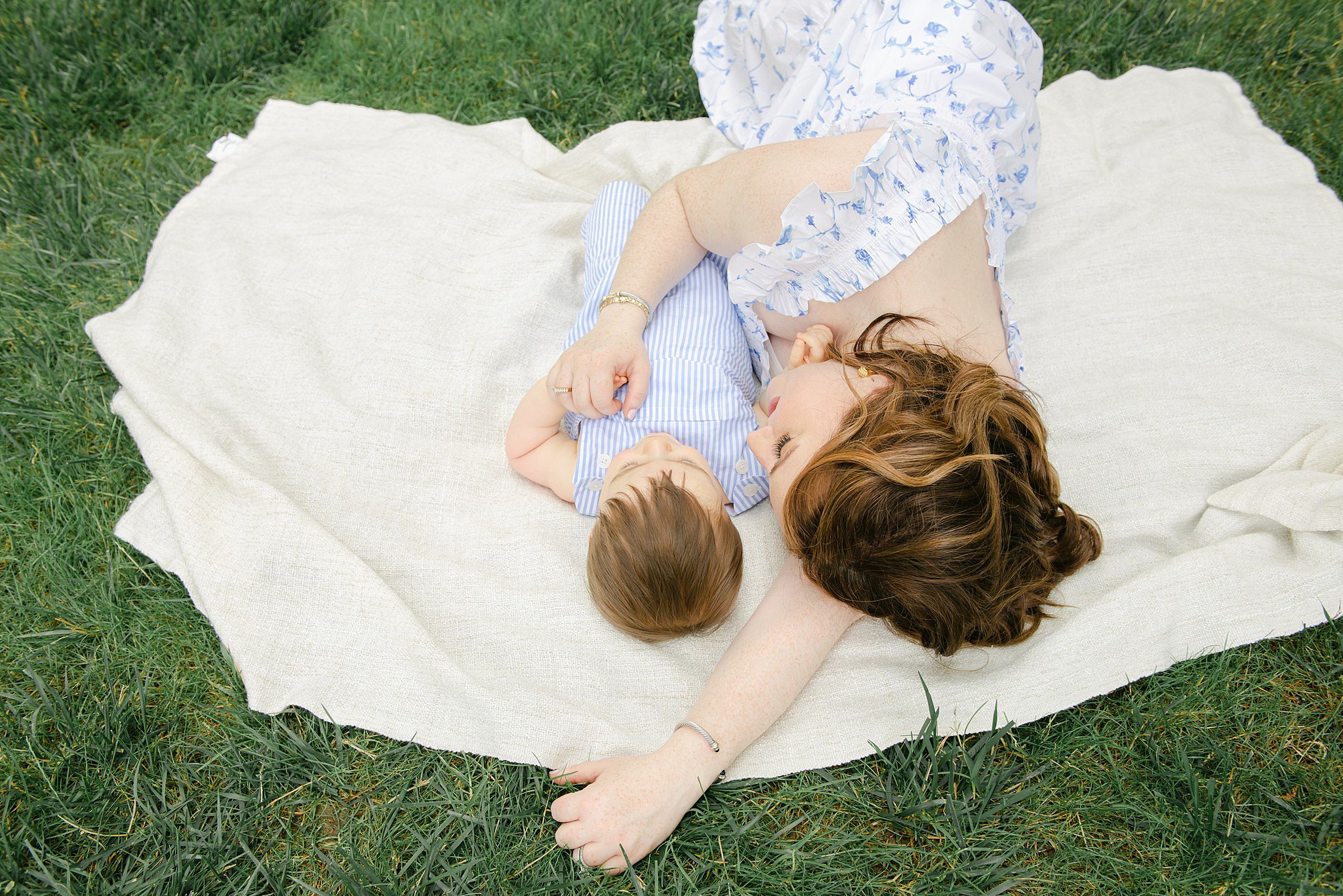 A mother in a blue dress lays on a picnic blanket in the grass with her toddler son in a blue striped onesie